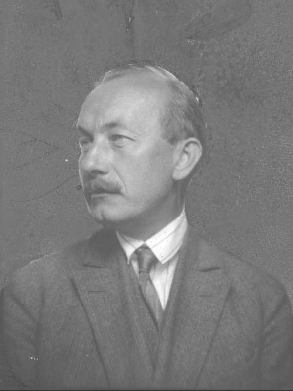 Józef Kostrzewski z profilu. Mężczyzna w średnim wieku, ma wysokie, łysiejące czoło, jasne oczy, wąsy, dołeczek w podbródku. Ubrany w garnitur, jasną koszulę i krawat.