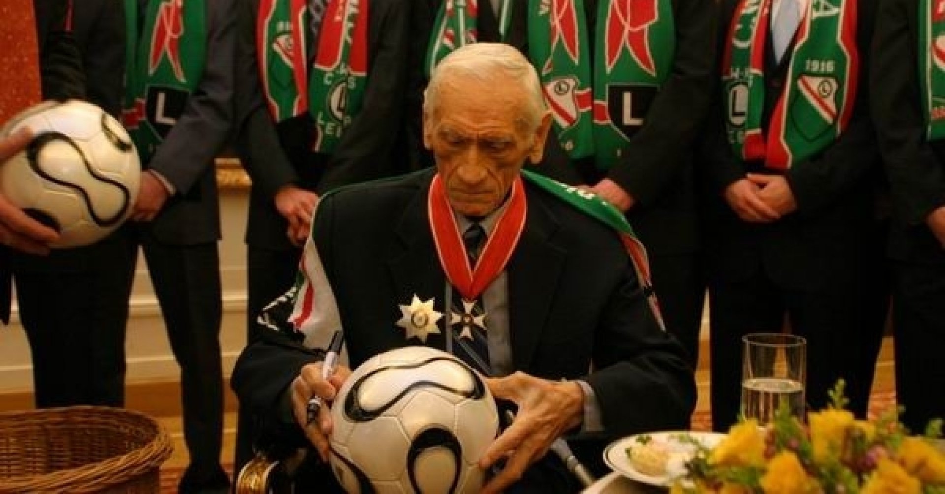 Kazimierz Górski siedzi przy stoliku z poczęstunkiem, na kolanach trzyma piłkę, w ręce czarny flamaster. Za trenerem stoi grupa młodych mężczyzn w szalikach klubu Legii Warszawa.