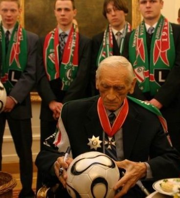 Kazimierz Górski siedzi przy stoliku z poczęstunkiem, na kolanach trzyma piłkę, w ręce czarny flamaster. Za trenerem stoi grupa młodych mężczyzn w szalikach klubu Legii Warszawa.