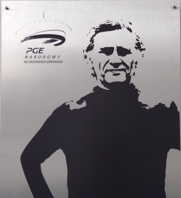 Grafika przedstawiająca sylwetkę Kazimierza Górskiego w czarnym golfie, z rękami wspartymi na biodrach. Po lewej stronie logo Stadionu Narodowego im. Kazimierza Górskiego.