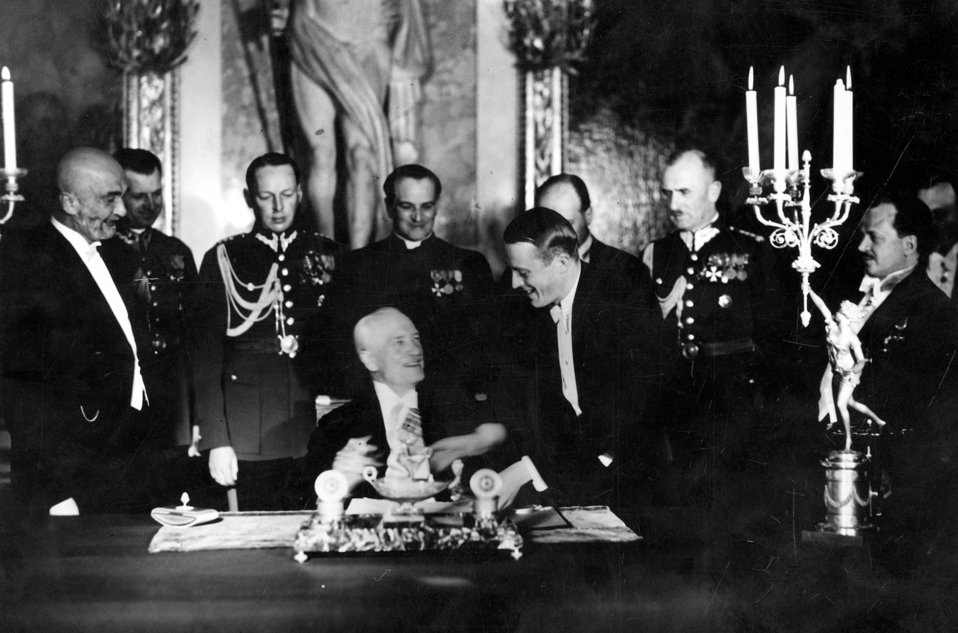 Siwy mężczyzna siedzi przy stole, wokół stoją mężczyźni we frakach i mundurach z odznaczeniami.