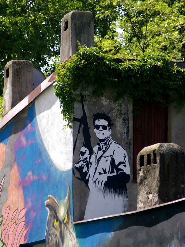 Mural namalowany na ścianie domu porośniętego dzikim winem przedstawia Zbigniewa Cybulskiego w płaszczu, ciemnych okularach i z karabinem w prawej ręce.