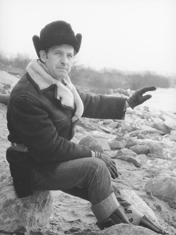 Andrzej Wajda w krótkim kożuszku, rękawiczkach i futrzanej czapce siedzi na kamieniu nad rzeką.