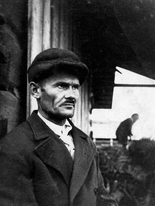 Mężczyzna o szupłej twarzy, ciemnych oczach, wyrazistych brwiach. Nosi krótkie wąsy. Ma na sobie płaszcz i czapkę. 