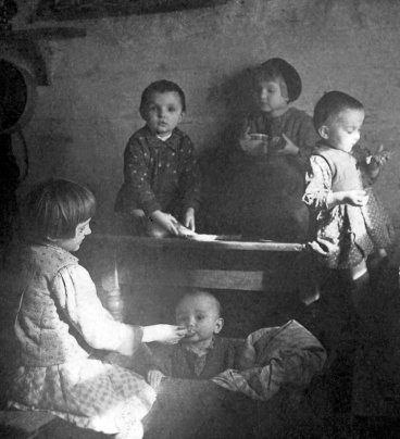 Pięcioro dzieci w oświetlonej słońcem izbie. Najstarsza dziewczynka karmi najmłodsze, może półroczne dziecko.