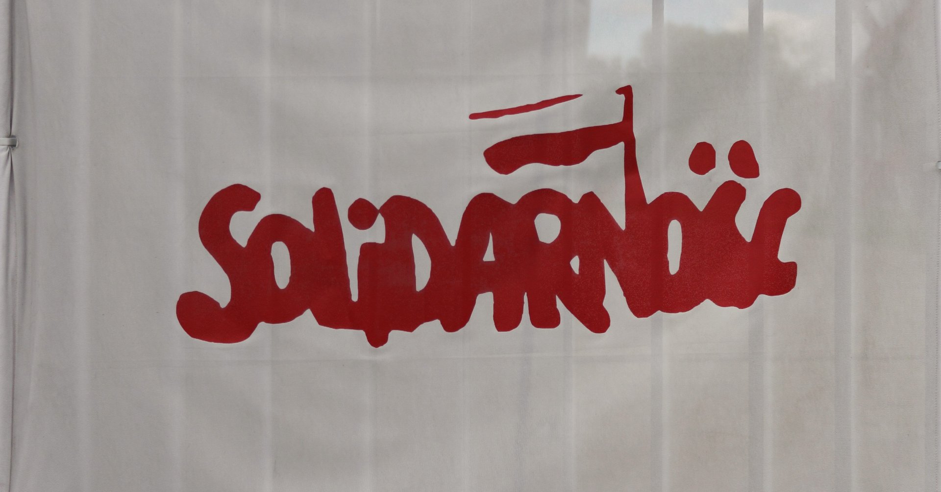 Na białym tle czerwony napis „Solidarność” z biało-czerwoną chorągiewką nad literą „N”.