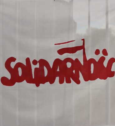 Na białym tle czerwony napis „Solidarność” z biało-czerwoną chorągiewką nad literą „N”.