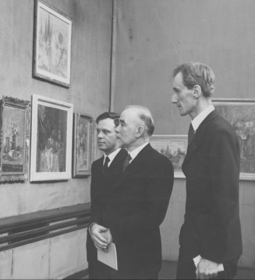 Trzej meżczyźni w białych koszulach i czarnych garniturach przyglądają się obrazom na wystawie. Józef Czapski wyróżnia się wysokim wzrostem.