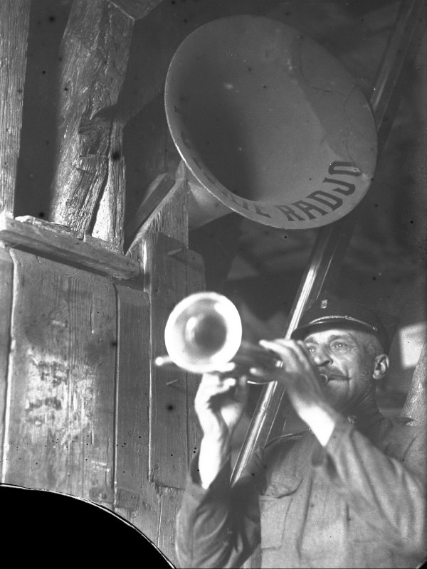 Mężczyzna w czapce i mundurze gra na trąbce. Nad nim umieszczona jest tuba z napisem „Polskie Radio”.