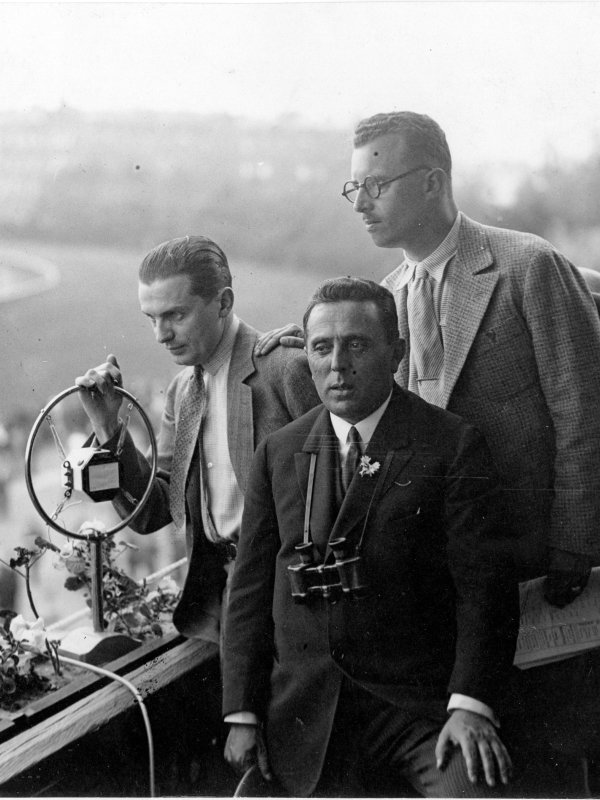 Na trybunie z widokiem na tor wyścigów stoi trzech młodych mężczyzn. Jeden zaciska rękę na mikrofonie radiowym. Drugi ma na szyi lornetkę. Trzeci obserwuje tor.