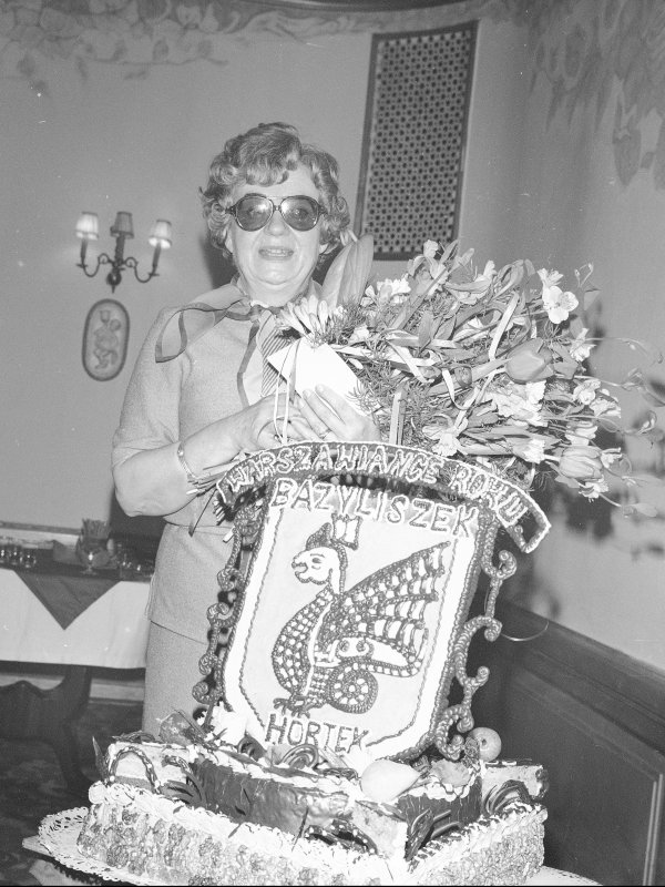Tęższa kobieta w średnim wieku, z krótkimi, kręconymi włosami, w ciemnych okularach, trzyma w rękach bukiet kwiatów. Obok stoi tort.