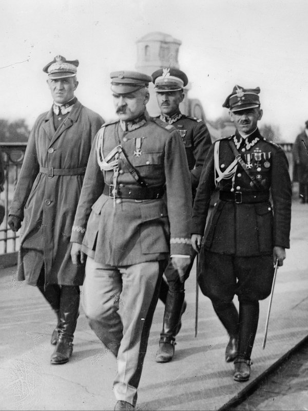 Józef Piłsudski na przedzie, za nim trzech oficerów. Wszyscy ubrani w mundury idą przez most.