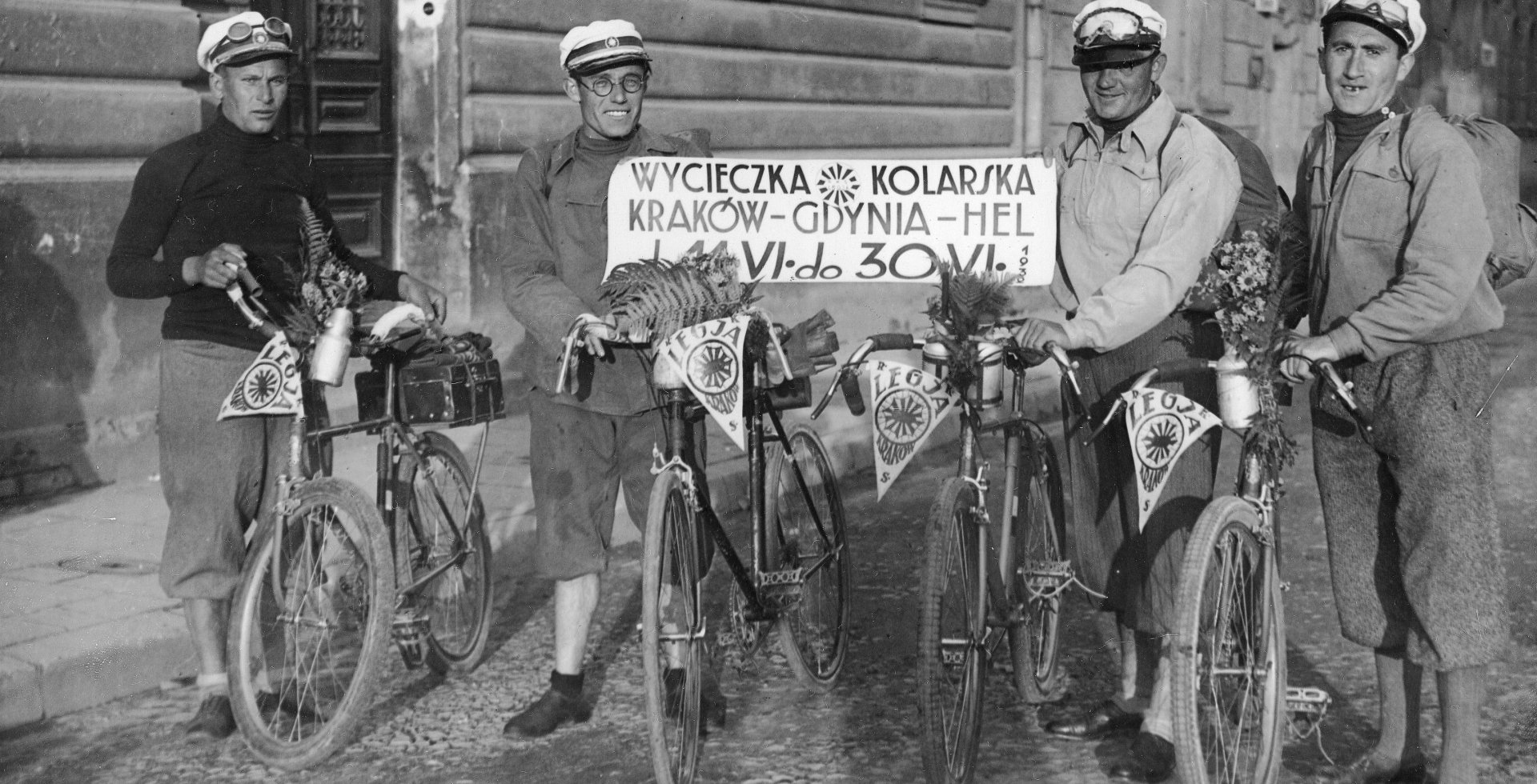 Czarno-biała fotografia przedstawia czterech mężczyzn pozujących z rowerami na miejskiej ulicy, w okresie międzywojennym.