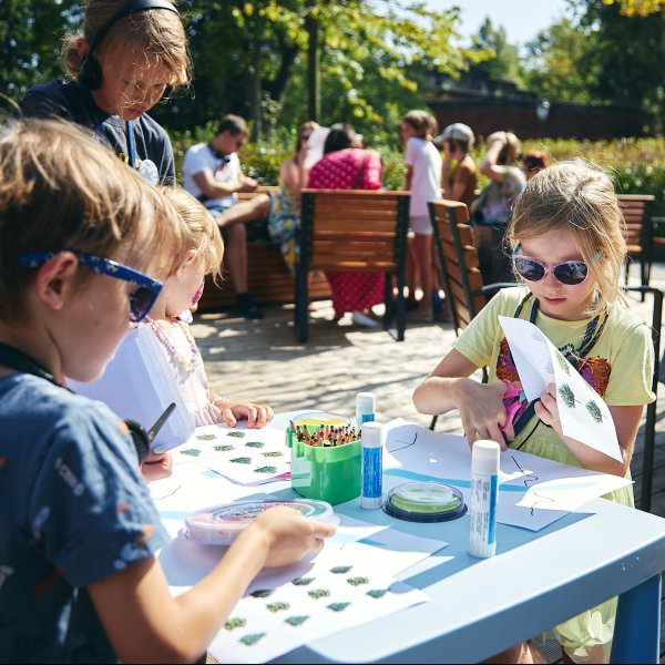 Zdjęcie z góry przedstawia troje dzieci siedzących przy jasnoniebieskim stoliku. Dzieci rysują kredkami na kartkach papieru. Na stole leżą kolorowe poduszki z tuszem do stempli, kredki w pojemniku, nożyczki oraz kartki z rysunkami małych choinek.