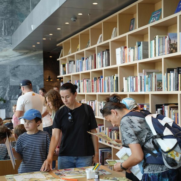 Zdjęcie przedstawia wnętrze biblioteki. Kobieta i chłopiec w niebieskiej czapce oglądają książki przy drewnianym stole. Druga kobieta z plecakiem przegląda publikacje. W tle widać wielki regał pełen książek oraz innych odwiedzających.