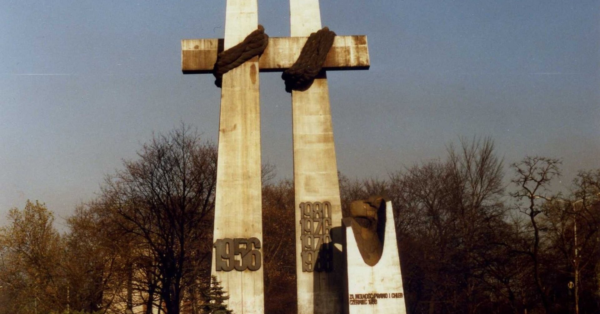 Dwa wysokie krzyże połaczone ze sobą ramionami. Raamiona przewiązane grubym sznurem. Na dole lewego krzyża data: 1956. Na dole prawego krzyża daty: 1968, 1970, 1876, 1980.