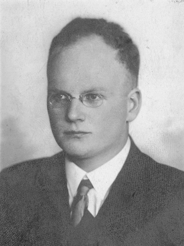 Młody mężczyzna o wysokim czole, inteligentnym wyrazie twarzy, w okularach.  Ma na sobie garnitur, białą koszulę i krawat.