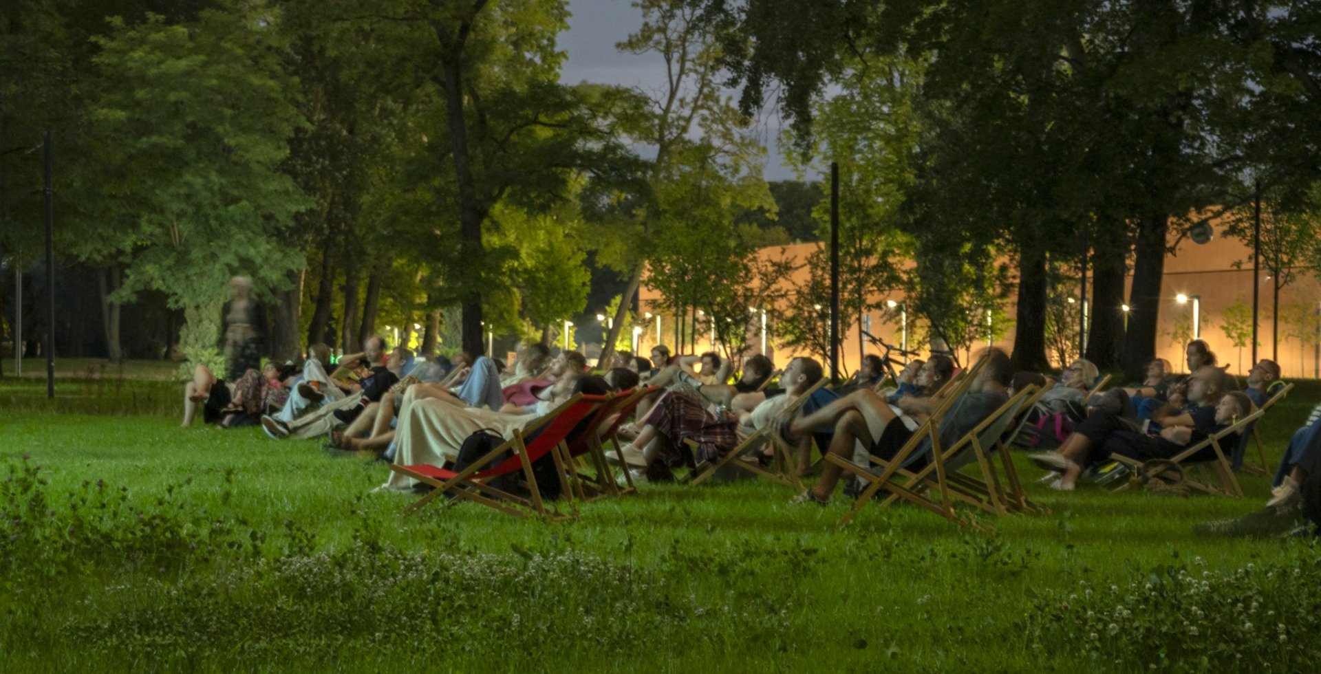 Widok z boku na publiczność kina plenerowego. Dziesiątki osób siedzą na rozstawionych na trawniku leżakach. W tle widać gęste drzewa oraz oświetlony budynek, a cała scena tonie w wieczornym półmroku.