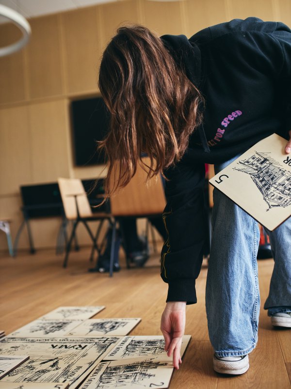 Bliskie ujęcie drewnianej tabliczki z wygrawerowanym rysunkiem bramy miejskiej i napisem BRAMA x5. W tle widoczne są inne materiały edukacyjne, w tym kartka z napisem „Cech kowali” oraz rozmyte sylwetki osób biorących udział w zajęciach historycznych.