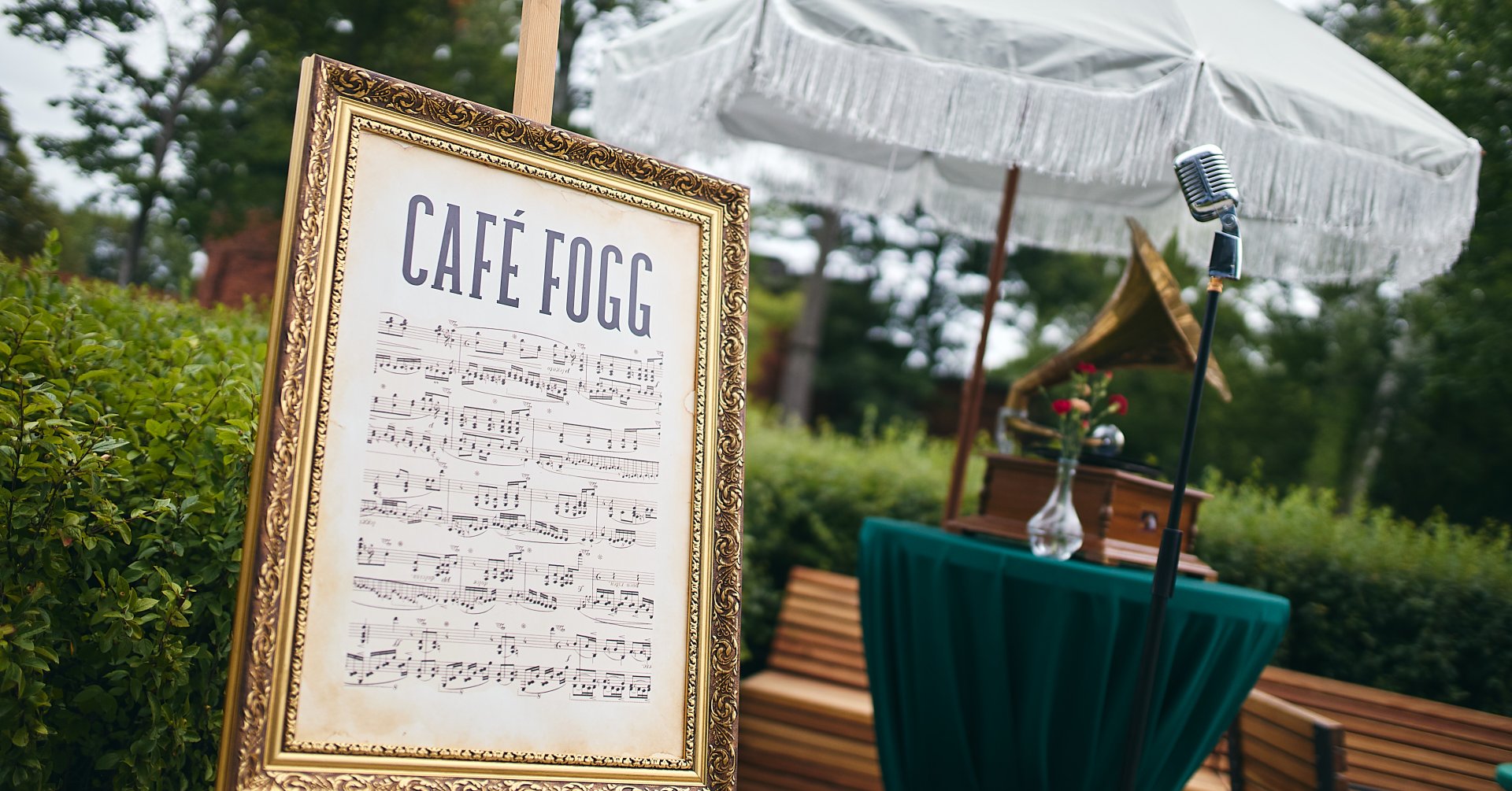 Pod białym parasolem w parku stoi kilka ławek, stolik z gramofonem. W złotej ramie ustawionej na sztalugach napis „Caffe Fogg”.