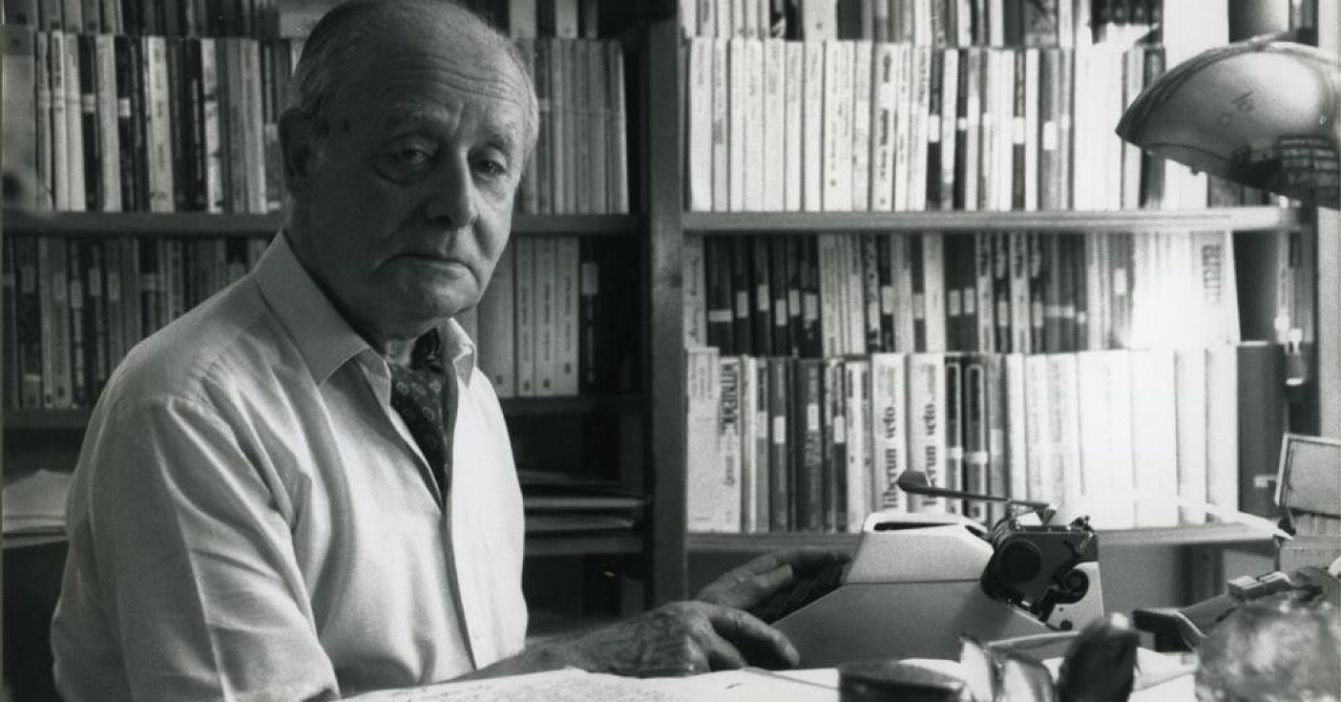 Jerzy Giedroyc w białej koszuli i fularze siedzi przy biurku z maszyną do pisania. W tle półki z mnóstwem książek.