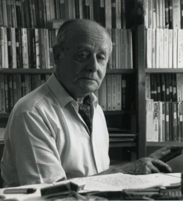Jerzy Giedroyc w białej koszuli i fularze siedzi przy biurku z maszyną do pisania. W tle półki z mnóstwem książek.