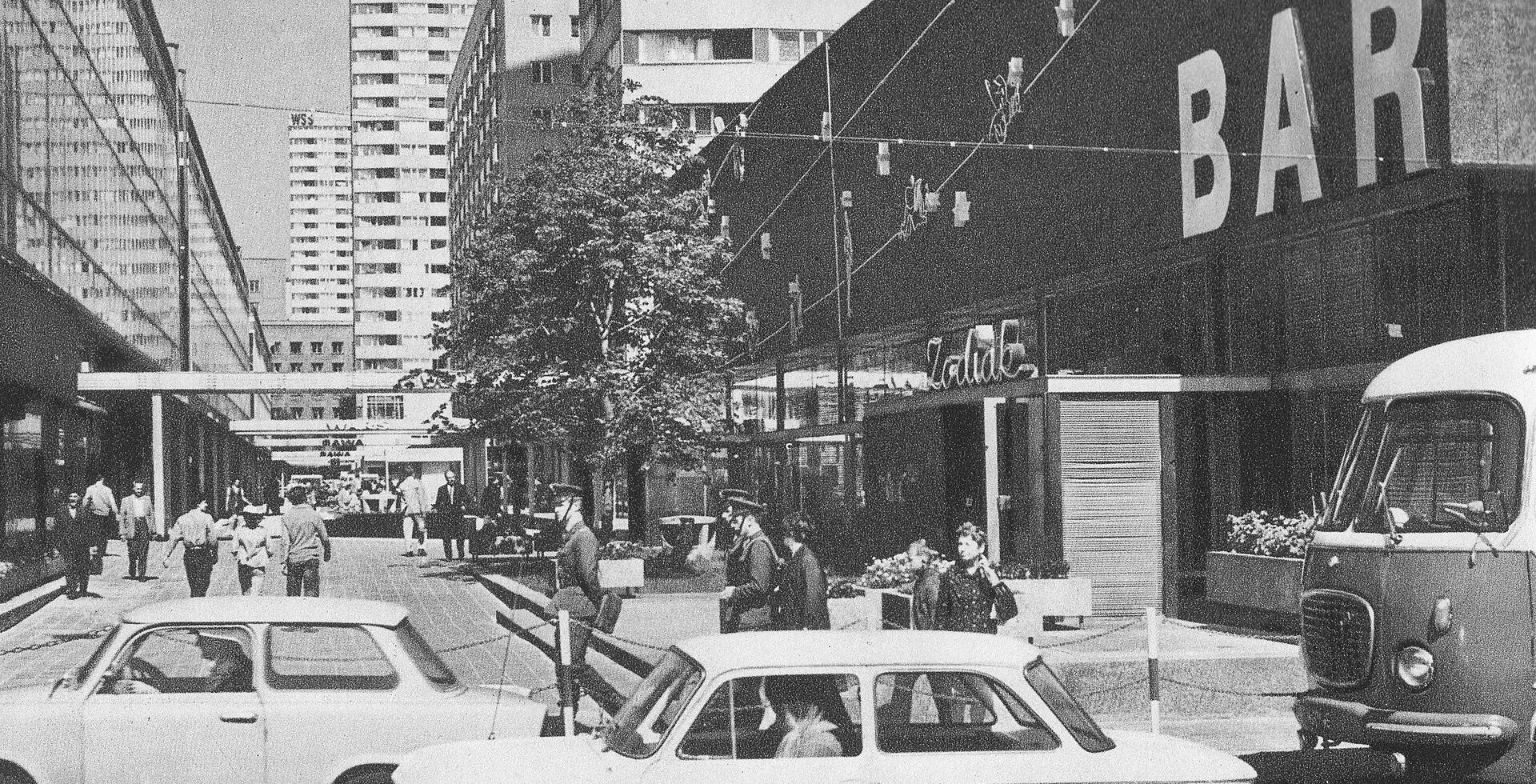 Bar Zodiak w Warszawie w latach 70. XX wieku. Na pierwszym planie samochody i przechodnie, w tle modernistyczne budynki centrum miasta. Fotografia ukazuje atmosferę miejskiego życia oraz architekturę i ruch uliczny epoki PRL.