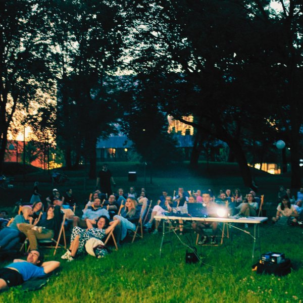 Kino plenerowe w parku o zmierzchu. Widzowie siedzą na leżakach i kocach rozłożonych na trawie przed niewidocznym ekranem. Pośrodku stoi stół z projektorem. W tle widać ciemne sylwetki drzew i nowoczesny budynek rozświetlony od wewnątrz.