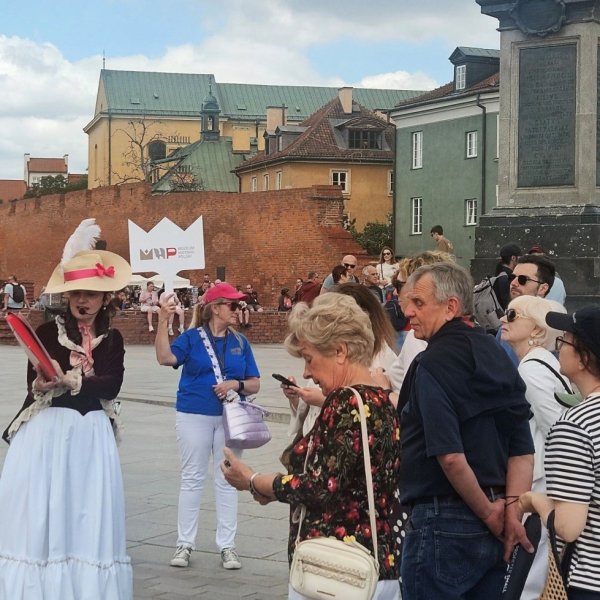 Przewodnik w stroju z epoki i kobieta z białym logo MHP oprowadzają grupę turystów po placu Zamkowym w Warszawie. W tle widoczna Kolumna Zygmunta, mury obronne Starego Miasta oraz kolorowe kamienice pod lekko zachmurzonym niebem.