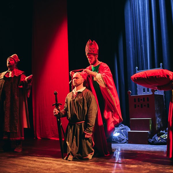 Aktorzy na scenie teatralnej. Klęczący mężczyzna w ciemnej szacie i z mieczem w dłoni jest koronowany przez postać w stroju biskupa. Obok stoją dwaj inni aktorzy w stylizowanych, dawnych strojach.