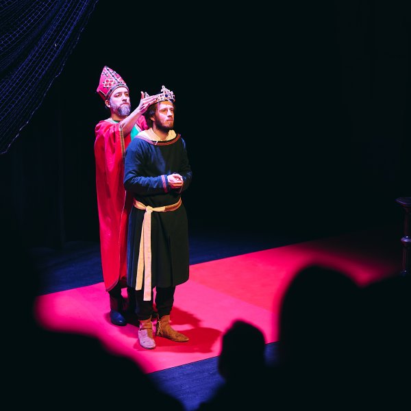 Scena teatralna w ciemnym otoczeniu. Postać w stroju biskupa nakłada koronę na głowę stojącego przed nią mężczyzny w ciemnej szacie z jasnym pasem. Aktorzy stoją na czerwonym podłożu, a w tle widać fragment siatki.