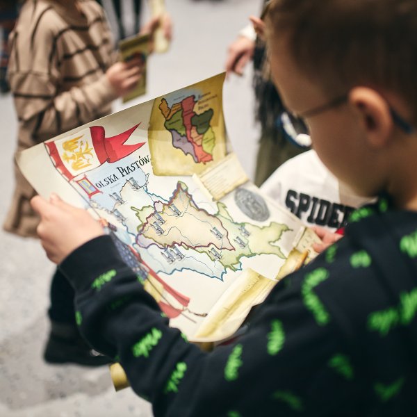 Chłopiec w okularach i ciemnej bluzie ogląda trzymaną w dłoniach kolorową, ilustrowaną mapę historyczną z napisem „Polska Piastów”. W tle widoczne są rozmyte sylwetki innych dzieci w jasnym pomieszczeniu.