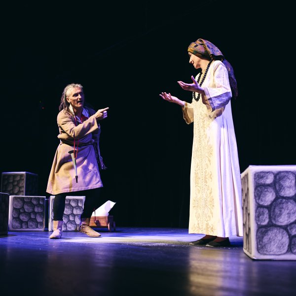 Scena teatralna na czarnym tle. Po lewej aktor w beżowym stroju żywo gestykuluje i wskazuje palcem na stojącą po prawej aktorkę w długiej, jasnej sukni i chustce na głowie. Na scenie leżą dekoracyjne kostki z motywem kamieni.