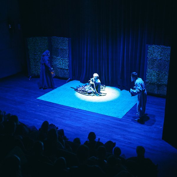 Scena teatralna oświetlona niebieskim światłem, widoczna z perspektywy widowni. Na środku siedzi postać w jasnym snopie światła. Po bokach stoją dwaj inni aktorzy – jeden w ciemnej szacie z kapturem, drugi w jasnym stroju.