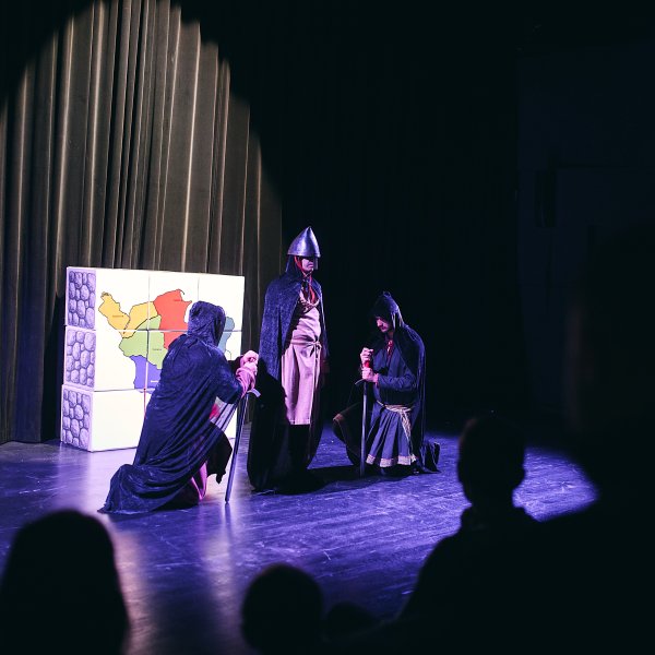 Scena teatralna oświetlona reflektorem. Trzej aktorzy w strojach historycznych: środkowy stoi w szpiczastym hełmie, a dwaj pozostali klęczą przed nim w ciemnych szatach z kapturami. W tle po lewej stronie widać dekorację z mapą.