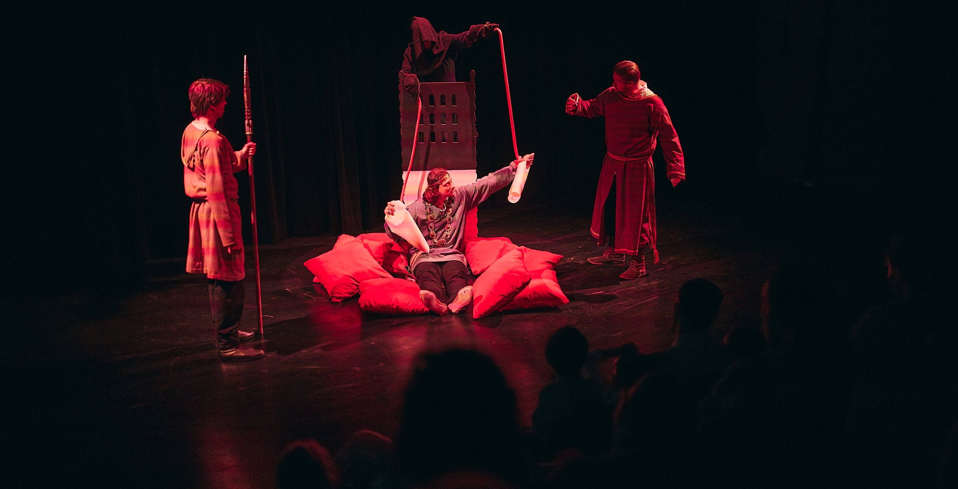 Aktorzy na scenie teatralnej oświetlonej czerwonym światłem. Na środku, wśród czerwonych poduszek, siedzi postać trzymająca zwoje papieru. Wokół stoją inni wykonawcy w dawnych strojach, w tym strażnik z włócznią i postać w kapturze.