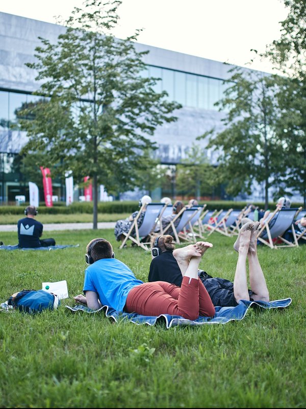 Zdjęcie przedstawia ludzi odpoczywających na trawniku przed nowoczesnym budynkiem Muzeum Historii Polski w Warszawie. Część osób leży na kocu w słuchawkach, inni siedzą w tle na leżakach. Wokół rosną zielone drzewa.