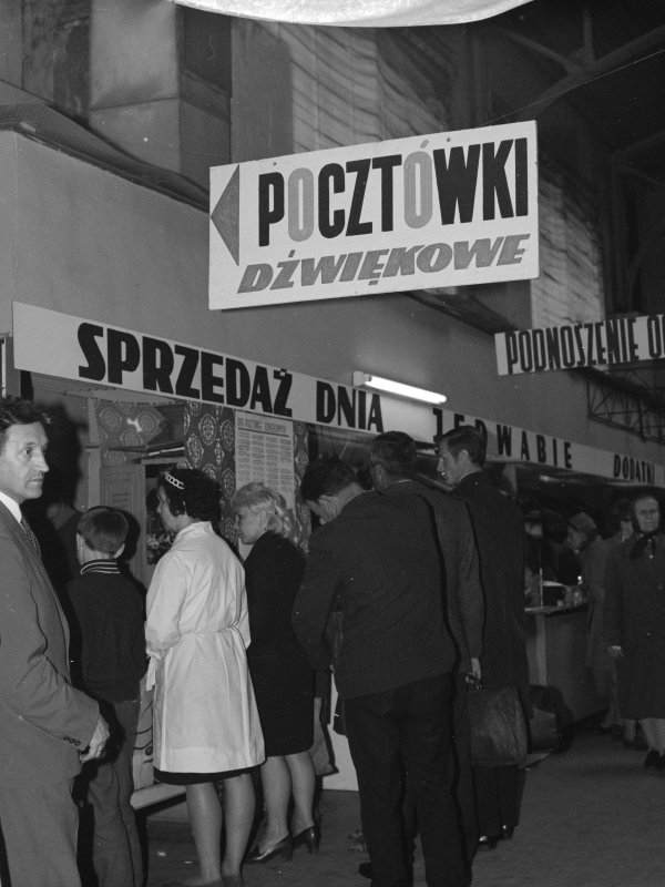 Czarno-białe zdjęcie przedstawia stoiska handlowe w czasach PRL. Widoczne są duże szyldy z napisami „Pocztówki dźwiękowe” oraz „Sprzedaż dnia...”. Przed ladami stoją klienci w kolejkach, a w tle widać tabliczkę z napisem „Podnoszenie oczek”.