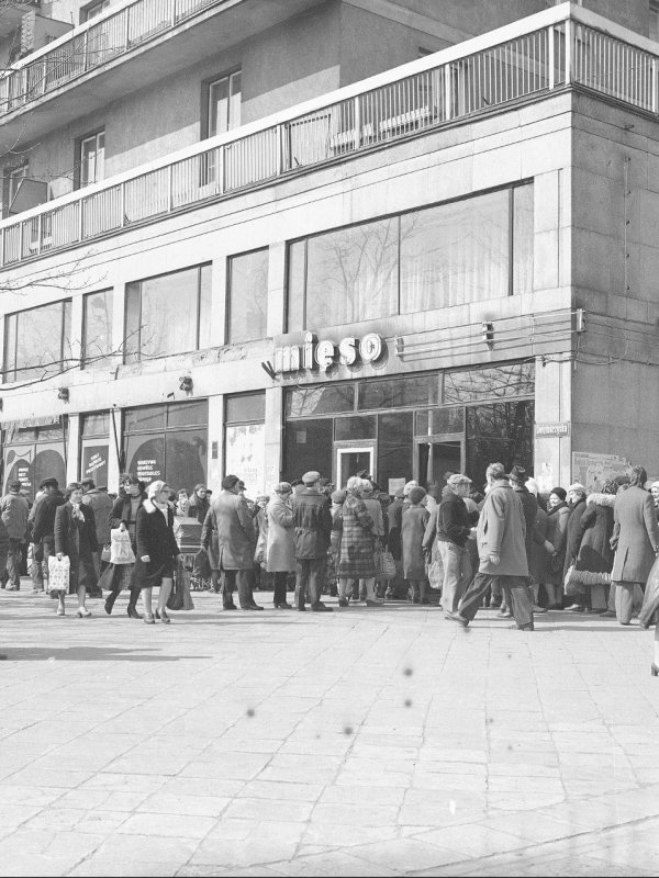 Czarno-białe zdjęcie przedstawia tłum ludzi stojących w kolejce przed sklepem z napisami „mięso” i „wędliny”. Sklep mieści się na parterze bloku z czasów PRL. Na dole kadru znajduje się czarny pasek Narodowego Archiwum Cyfrowego.