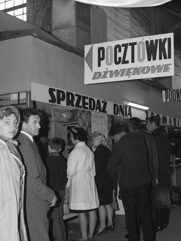 Czarno-białe zdjęcie z 1969 roku przedstawia kolejkę ludzi w hali targowej pod dużym szyldem z napisem „POCZTÓWKI DŹWIĘKOWE”. Kobiety i mężczyźni stoją tyłem, czekając na zakup przy ladzie, nad którą widnieje napis „SPRZEDAŻ DNIA...”.