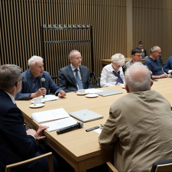 Zdjęcie przedstawia grupę osób siedzących wokół dużego, drewnianego stołu konferencyjnego podczas spotkania. Mężczyzna po lewej stronie mówi do mikrofonu. Na stole leżą dokumenty i laptopy. W tle widać nowoczesną ścianę z pionowych, drewnianych listew.