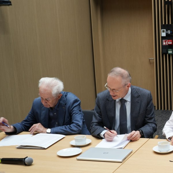 Zdjęcie przedstawia pięć osób siedzących w rzędzie przy drewnianym stole. Uczestnicy spotkania notują coś na leżących przed nimi dokumentach. Na stole stoją filiżanki i zielona butelka. W tle widać ścianę z pionowych, drewnianych listew.