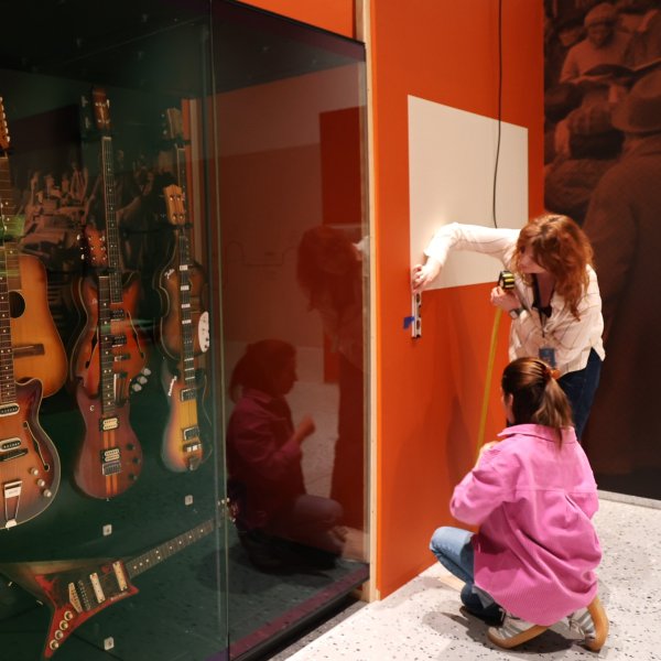Dwie kobiety pracują przy pomarańczowej ścianie ekspozycyjnej w muzeum. Obok nich stoi szklana gablota z gitarami elektrycznymi i basowymi. Po lewej stronie widoczny jest interaktywny ekran dotykowy z napisem „Dotknij”.