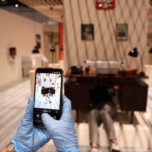 Dłonie w niebieskich rękawiczkach trzymają smartfon i robią zdjęcie scenie przed nimi. Na ekranie widać podgląd kadru z biurkiem. W rozmazanym tle znajduje się historyczna aranżacja gabinetu oraz osoba leżąca pod biurkiem.