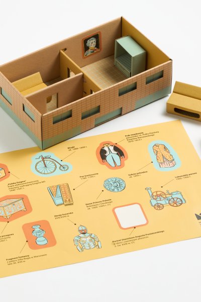 Zdjęcie przedstawia tekturowy model domku z mebelkami oraz żółtą planszę z ilustracjami zabytków, takimi jak bicykl czy zbroja husarska. Elementy edukacyjnego zestawu „Muzeum w pudełku” leżą na białym tle, tworząc przestrzenną makietę.