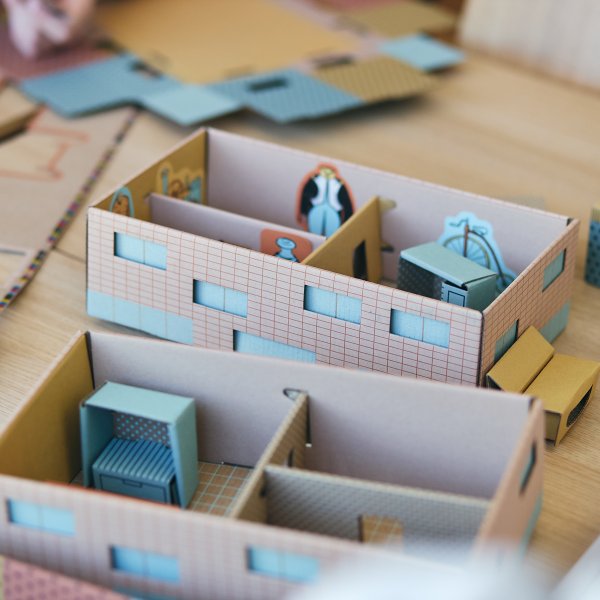 Zdjęcie przedstawia dwa tekturowe modele domków edukacyjnych ustawione na drewnianym stole. We wnętrzach pokoi widać miniaturowe mebelki oraz naklejki z zabytkami, m.in. bicyklem. Wokół leżą porozrzucane, wycięte elementy z kartonu.