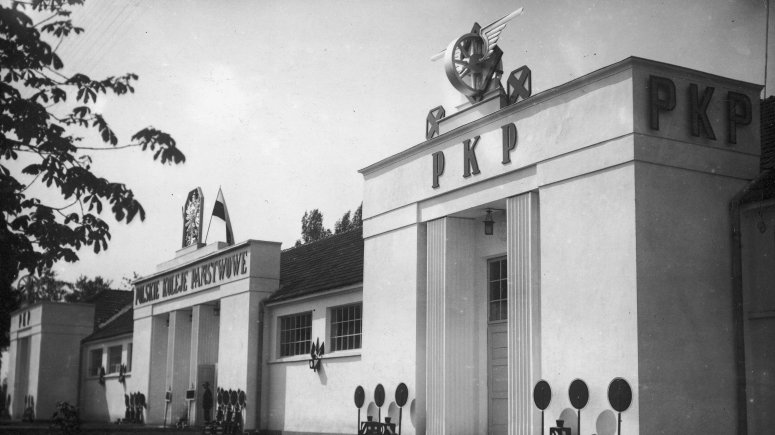 BIały budynek z kolumienkami. Na budynku widoczny napis „Polskie Koleje Państwowe”, polska flaga i godło.
