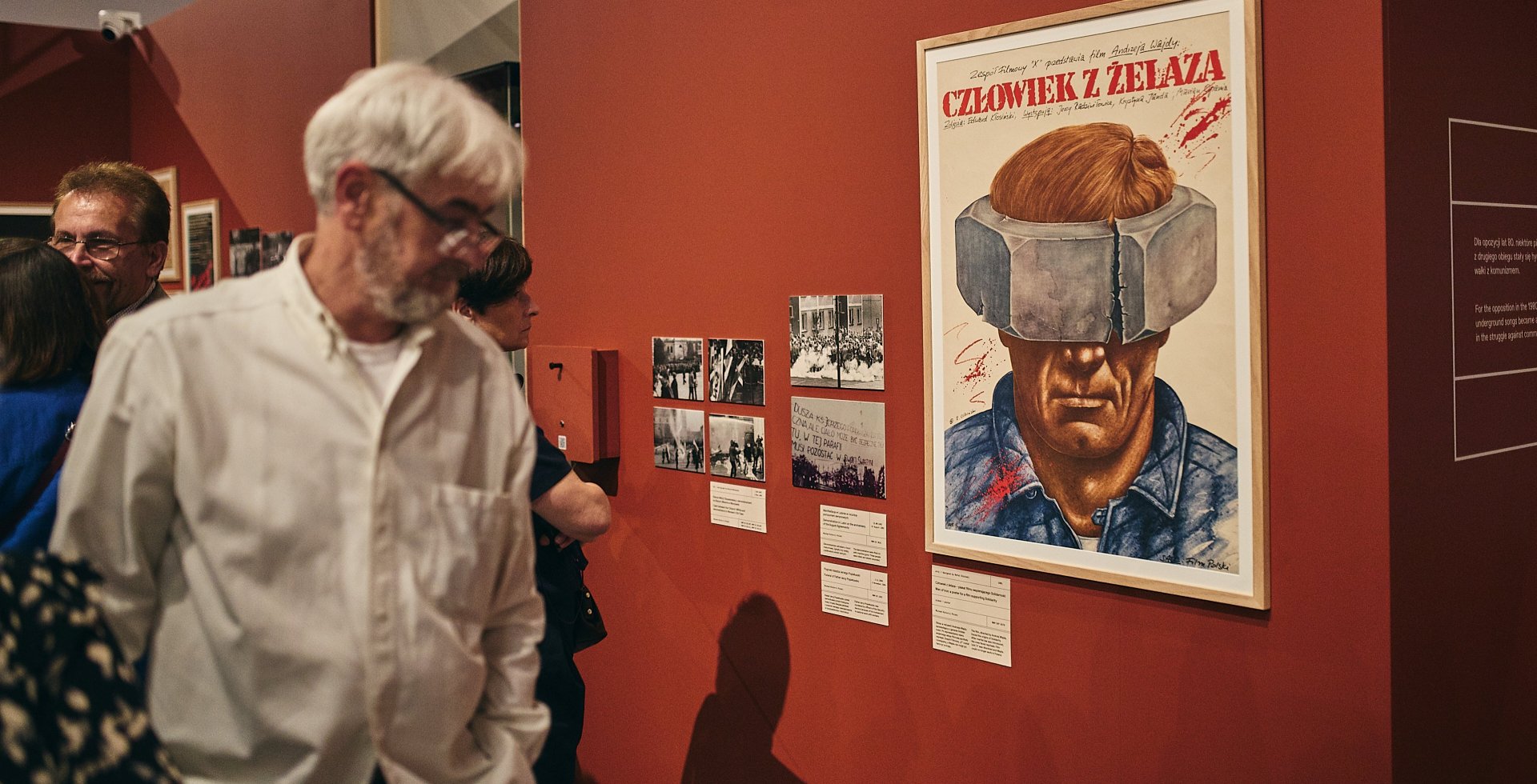 Zdjęcie przedstawia wystawę w muzeum na tle czerwonej ściany. Na pierwszym planie widać starszego mężczyznę w okularach i białej koszuli. Obok wisi plakat filmu „Człowiek z żelaza” autorstwa Rafała Olbińskiego oraz czarno-białe fotografie.