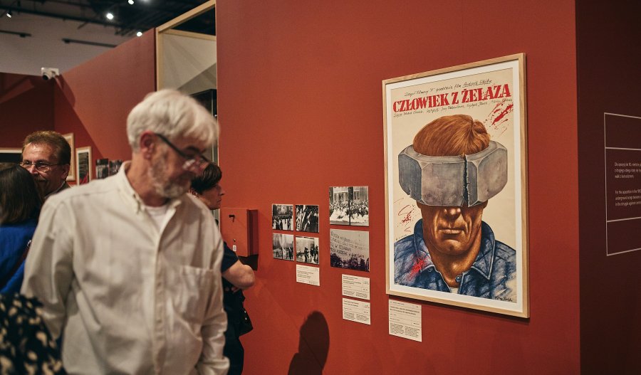Zdjęcie przedstawia wystawę w muzeum na tle czerwonej ściany. Na pierwszym planie widać starszego mężczyznę w okularach i białej koszuli. Obok wisi plakat filmu „Człowiek z żelaza” autorstwa Rafała Olbińskiego oraz czarno-białe fotografie.