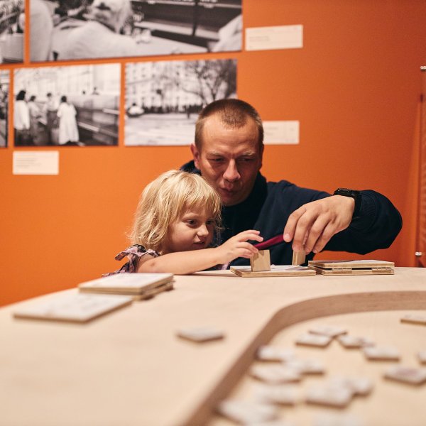 Mężczyzna z małą, jasnowłosą dziewczynką układają drewniane puzzle na stanowisku interaktywnym w muzeum. W tle widać jaskrawopomarańczową ścianę, na której wiszą czarno-białe, archiwalne fotografie.